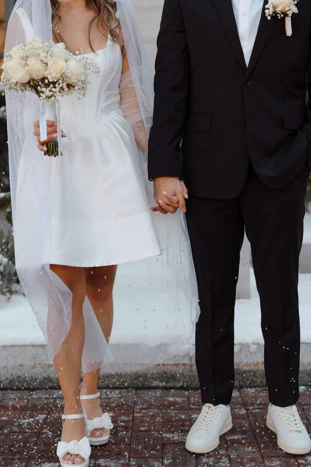 Close-up of bride and groom's matching white footwear with her short dress and his black suit visible.