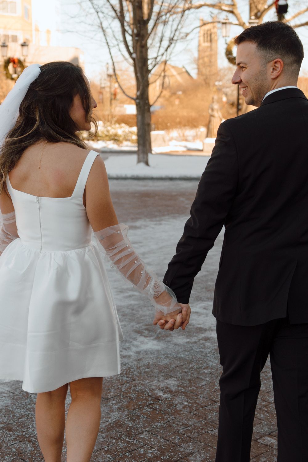 A couple walking together in winter, bride wearing short white dress and groom in black suit near a snowy sidewalk.