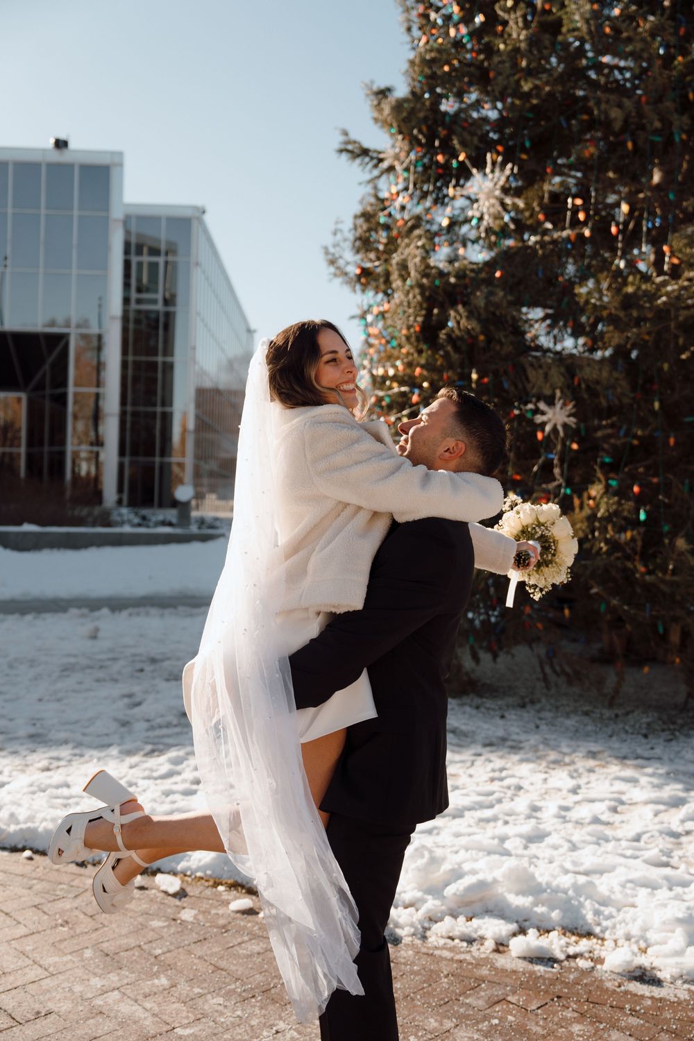 A joyful winter wedding moment with the couple embracing in front of a decorated Christmas tree in the snow.