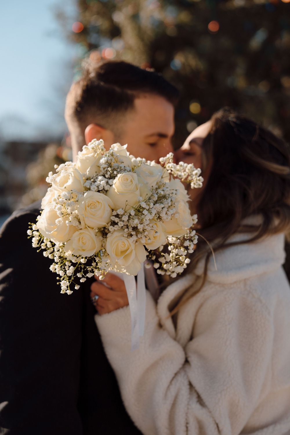 A romantic couple shares an intimate moment behind a beautiful white rose and baby's breath wedding bouquet.