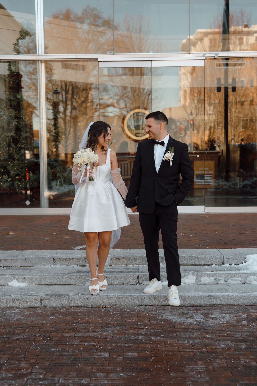 Bride in short dress and sneakers shares playful outdoor moments with groom near reflective building windows.