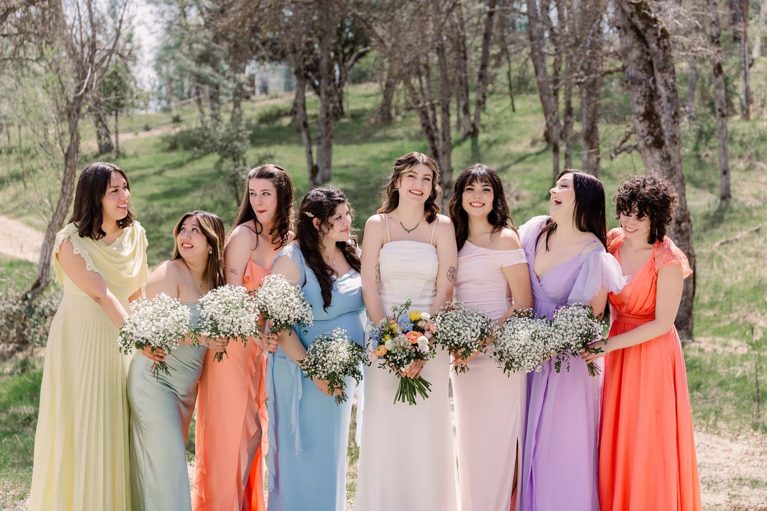 Bridesmaids wearing pastel dresses in different colors stand together at an outdoor wedding.