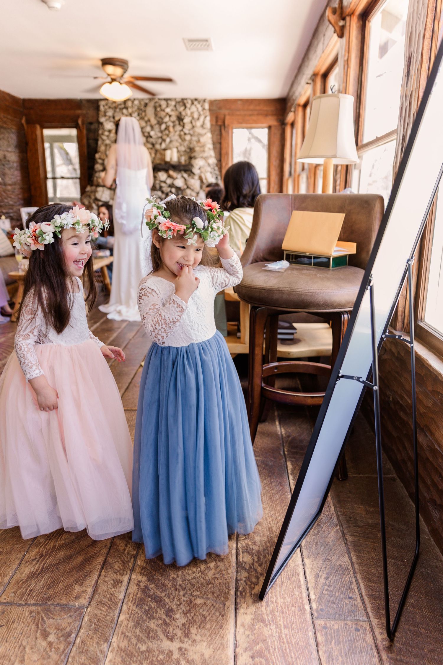 Bridesmaids in blue and pink dresses gathered around a mirror in a rustic getting-ready room.