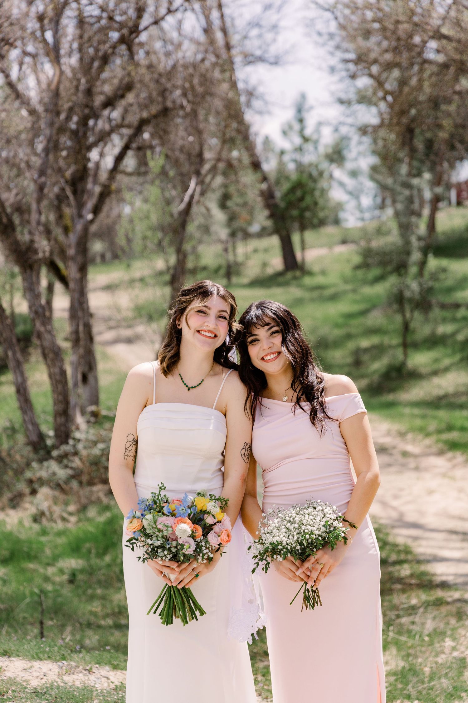 Two bridesmaids in pink dresses hold rustic wildflower bouquets in a sunny outdoor setting.