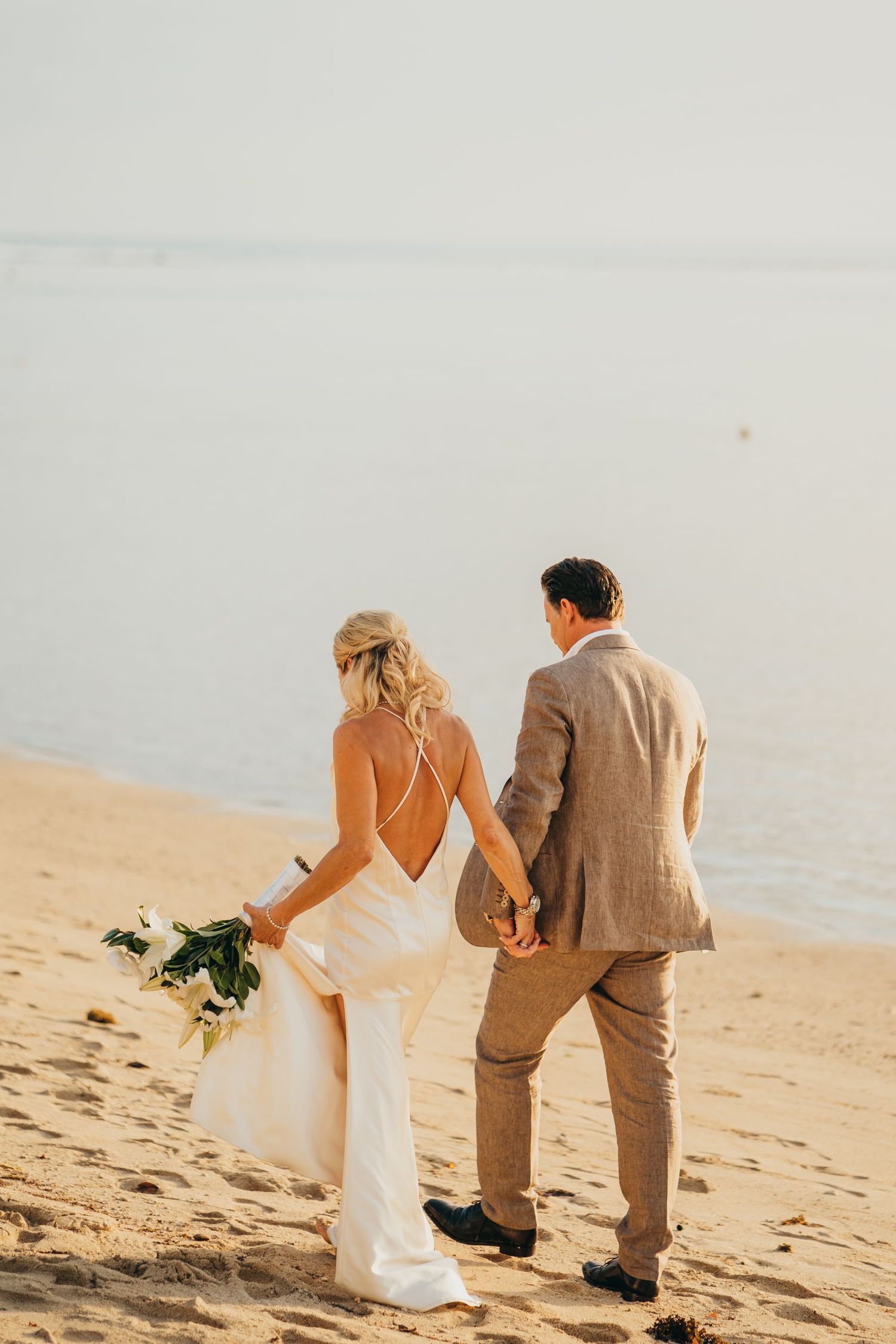 Rückenansicht eines Brautpaars am Strand. Die Braut trägt ein rückenfreies Brautkleid mit feinen Trägern.