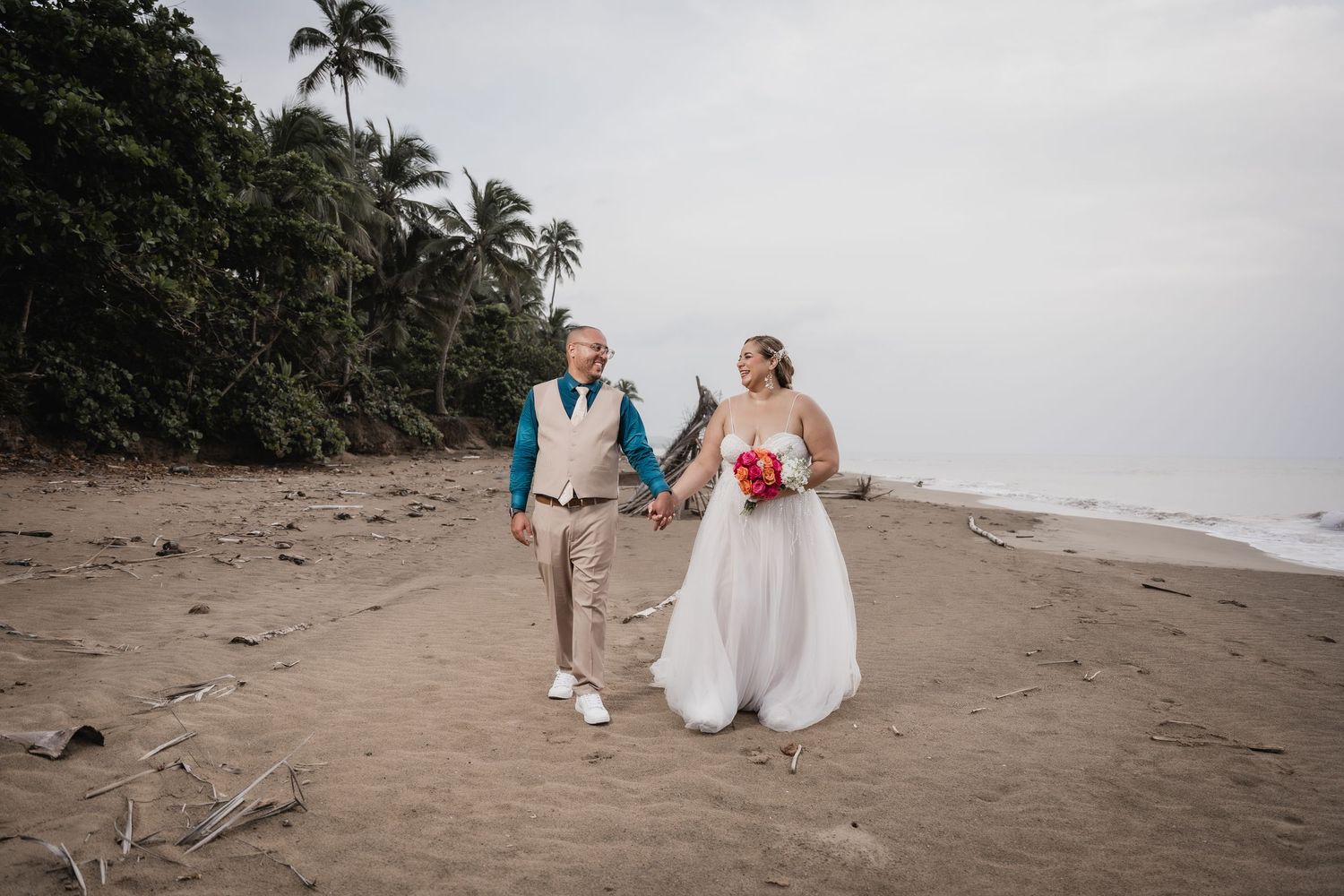 Beachside wedding couple walking along tropical shore in casual attire.