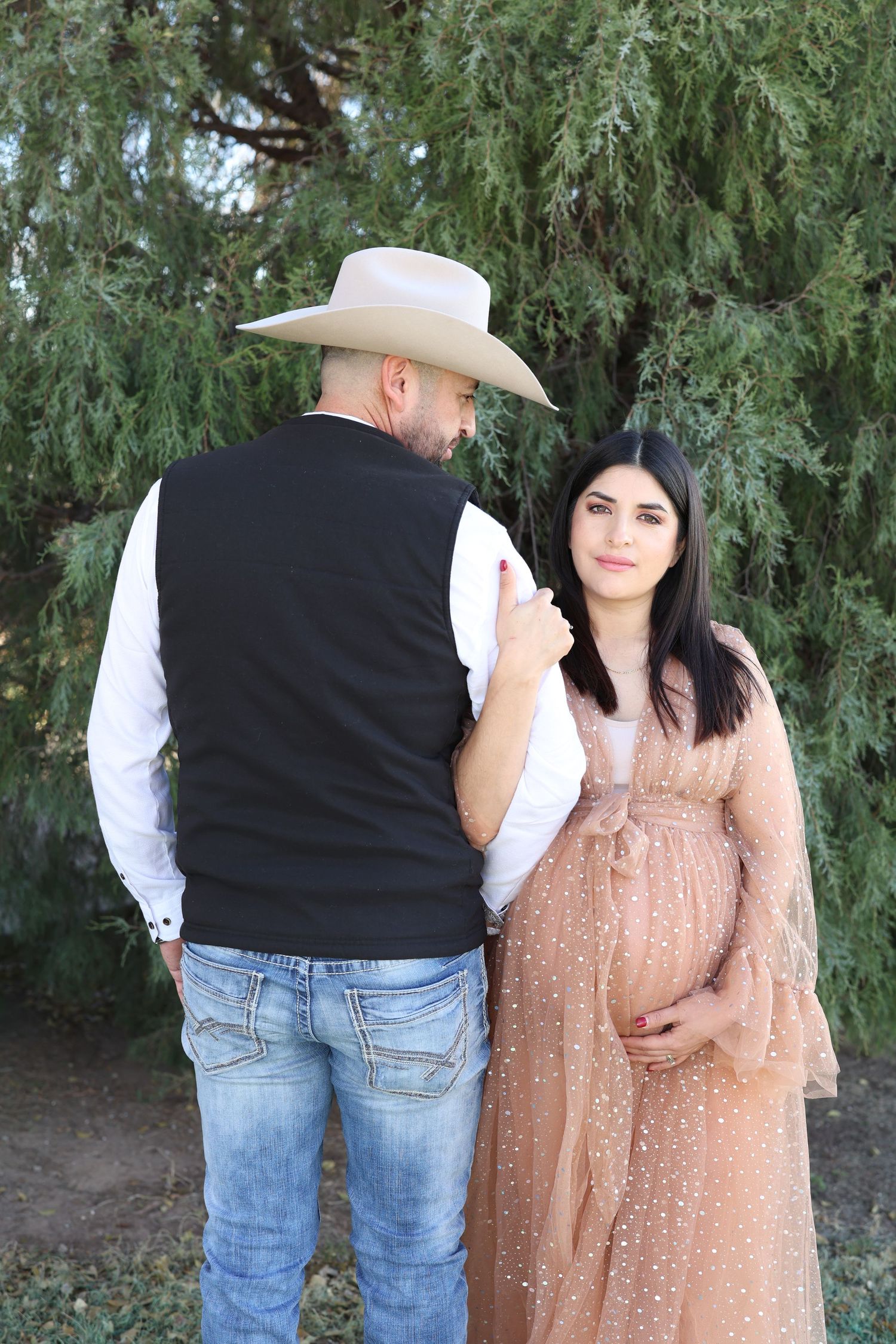 A couple in western attire sharing a tender moment against a backdrop of green trees.