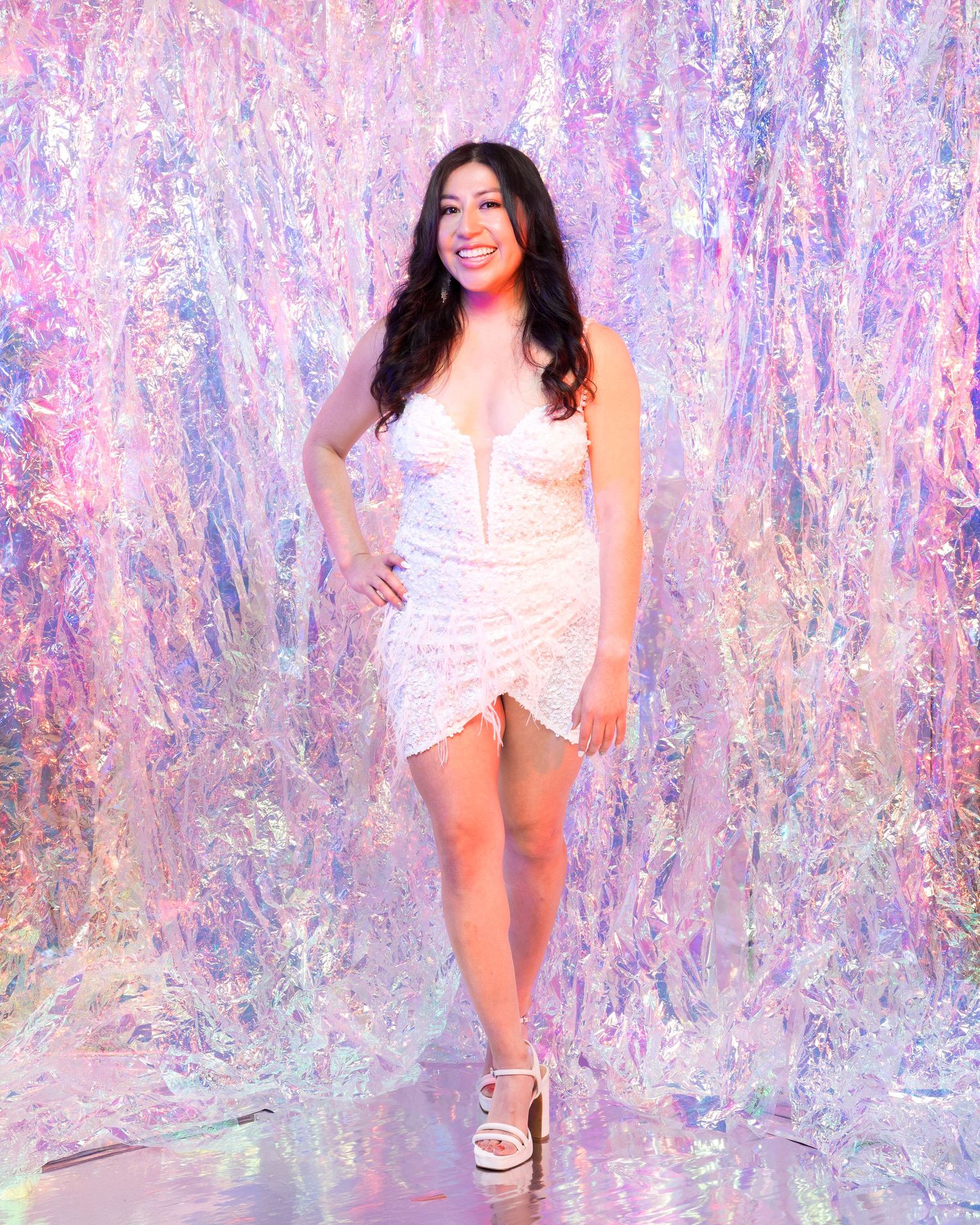 Woman in white dress poses against iridescent backdrop with pink and purple tones.
