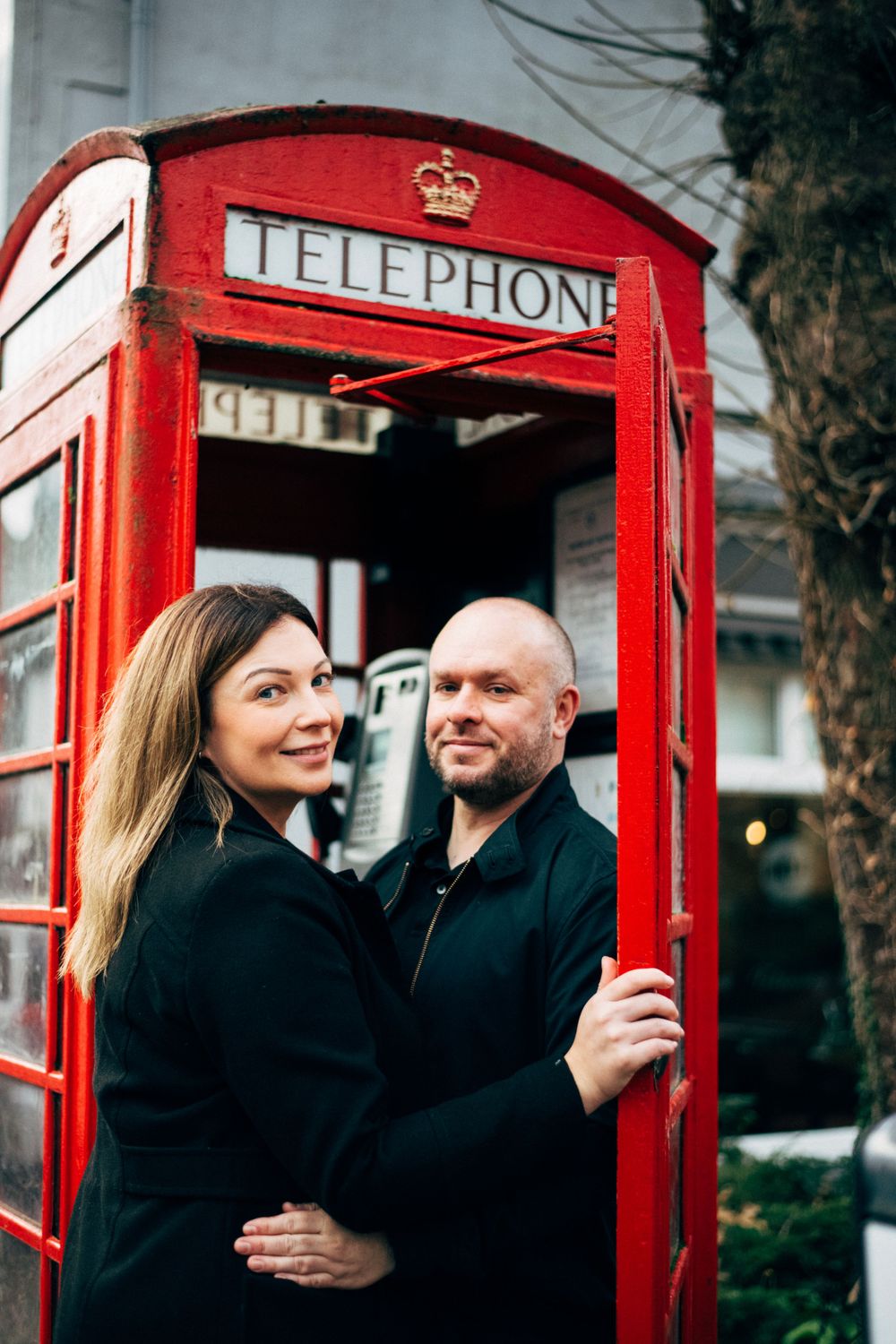 A couple poses together inside a classic red British phone booth.