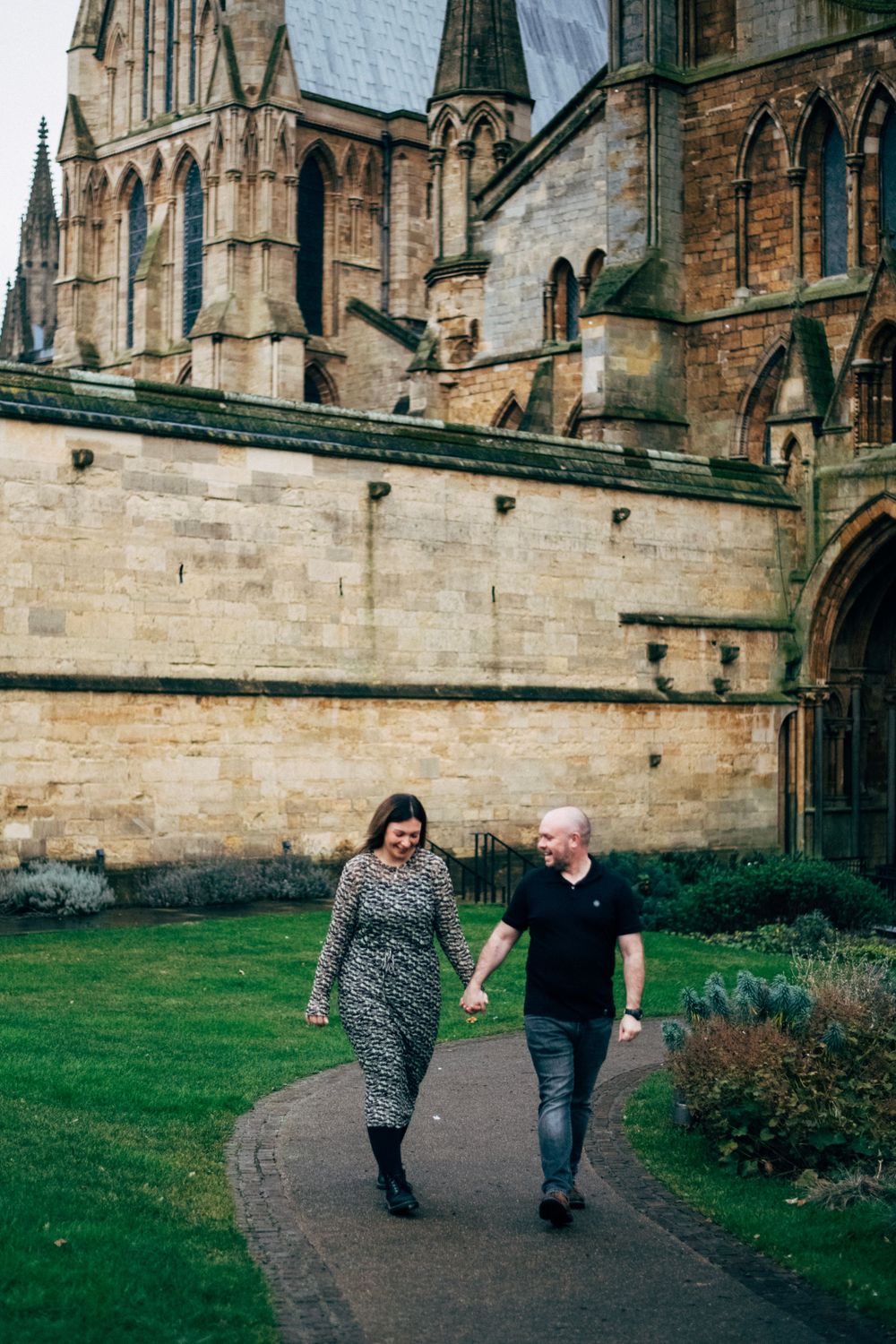A couple walks hand in hand along a path by a gothic cathedral.