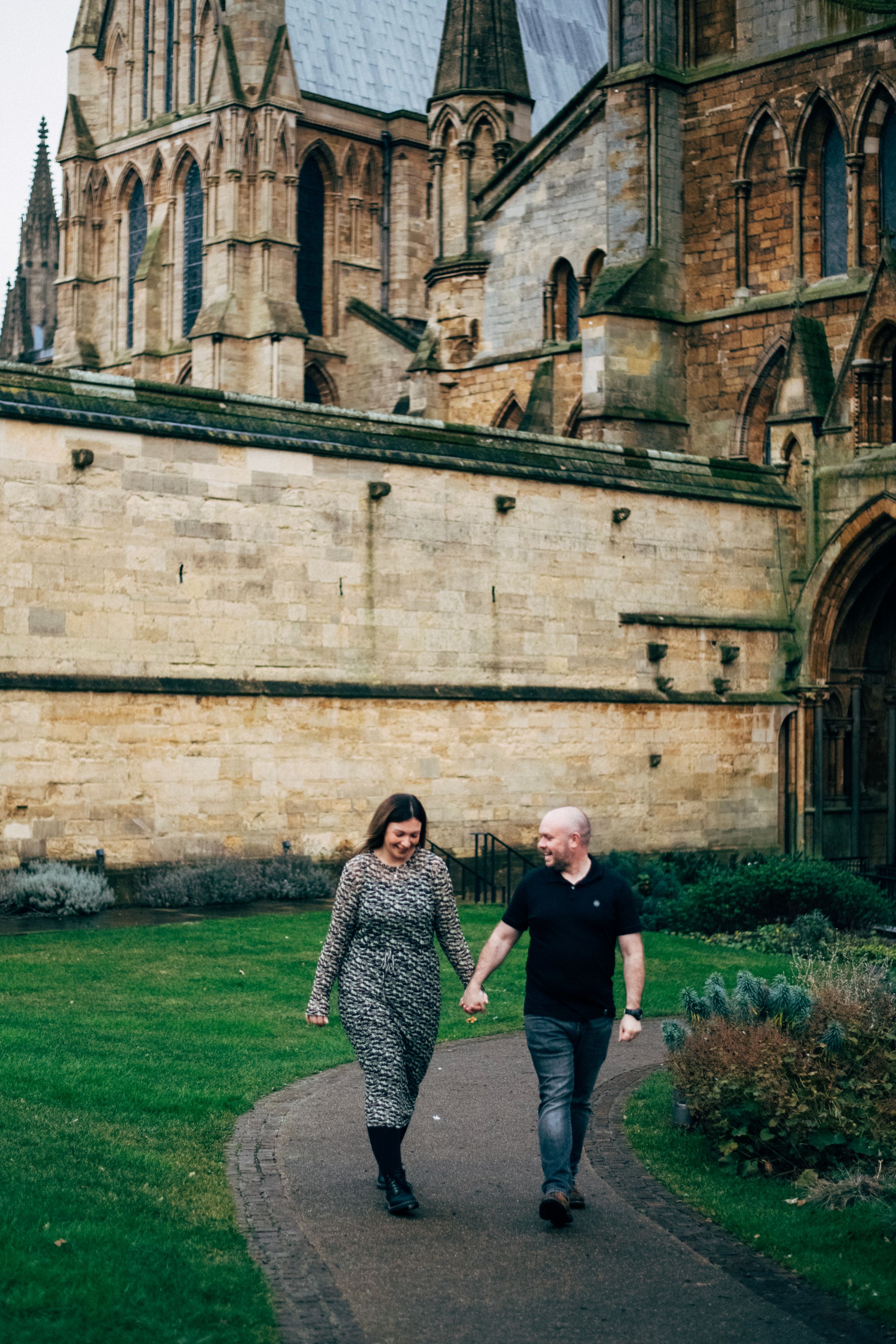 A couple walks hand in hand along a path by a gothic cathedral.