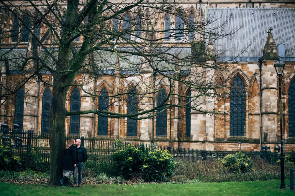 A couple stands among bare winter trees with cathedral architecture in the background.