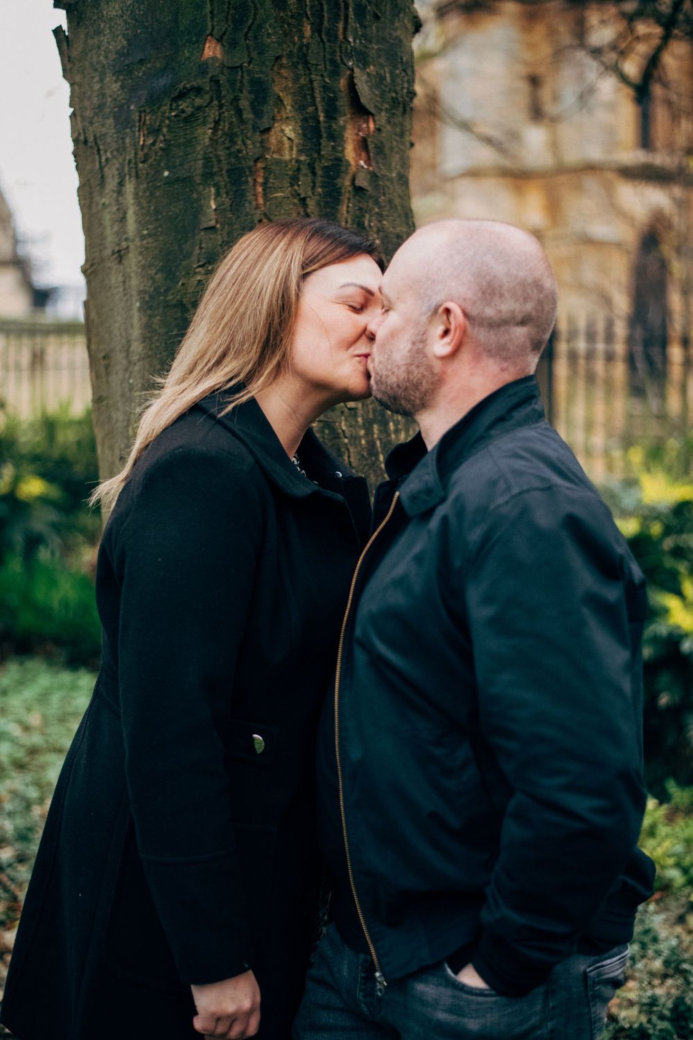 A couple shares an intimate kiss near a tree with cathedral architecture visible behind them.