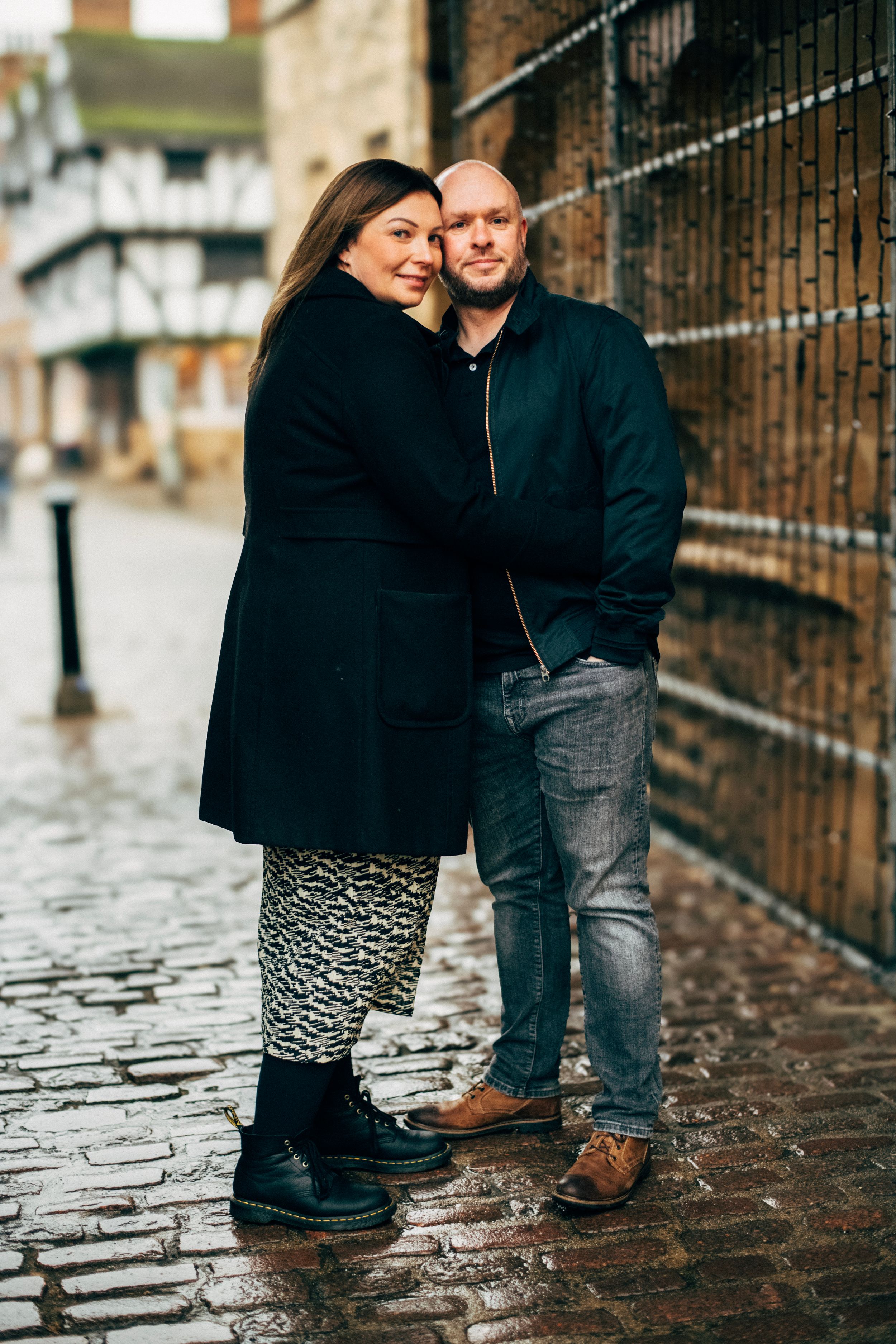 A couple stands close together on a cobblestone street against a brick wall on a rainy day.