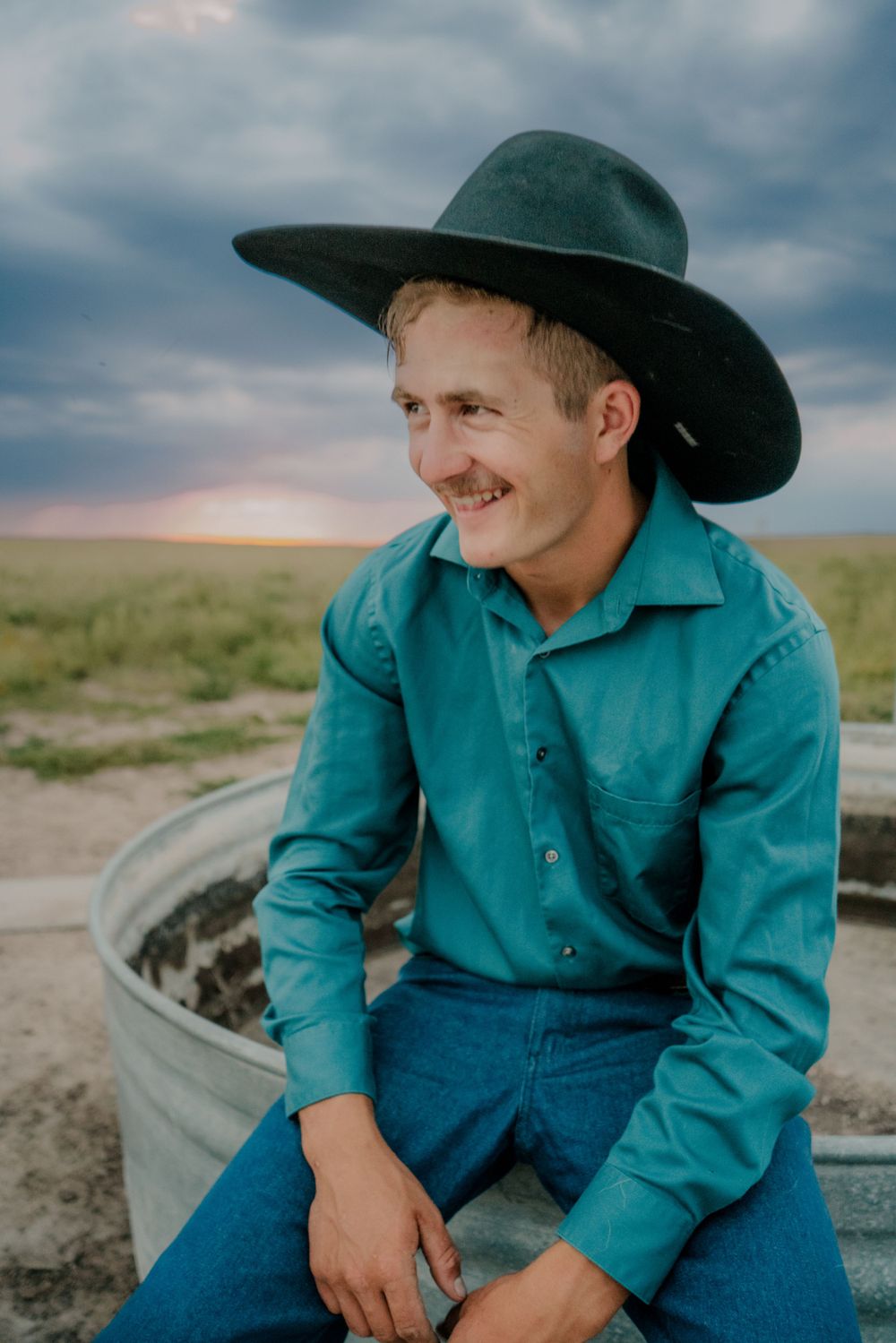 A cowboy sitting on a stock tank laughing while looking into the future with deep storm clouds in the background