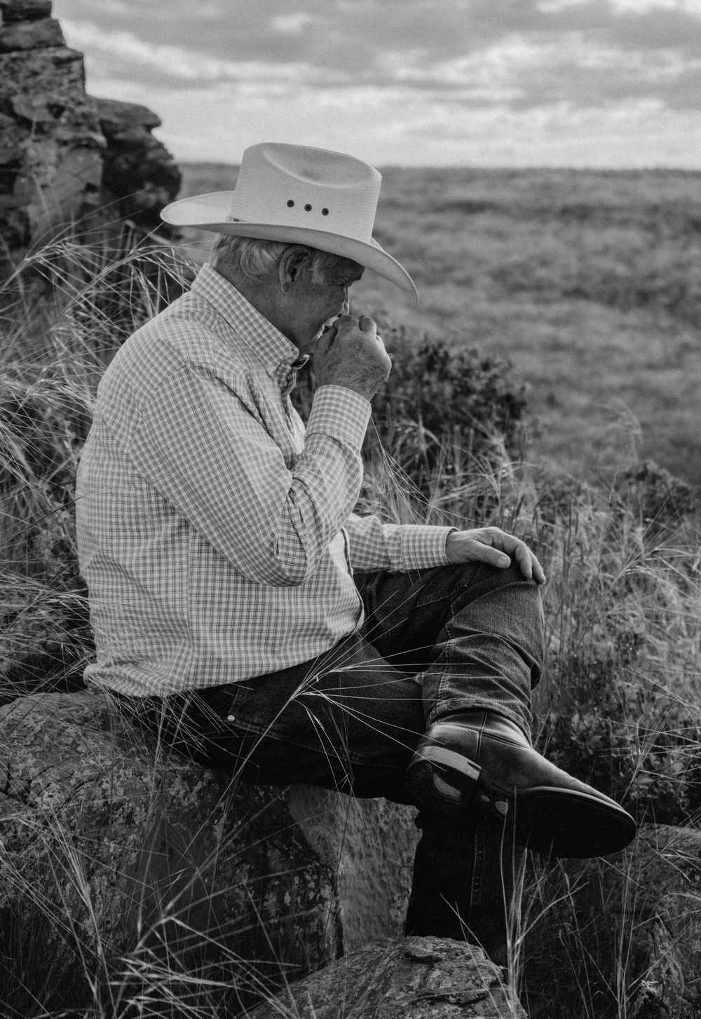 Black and white image with an elderly man in a cowboy hat sitting on a rock chewing on a pice of grass while looking into the distance