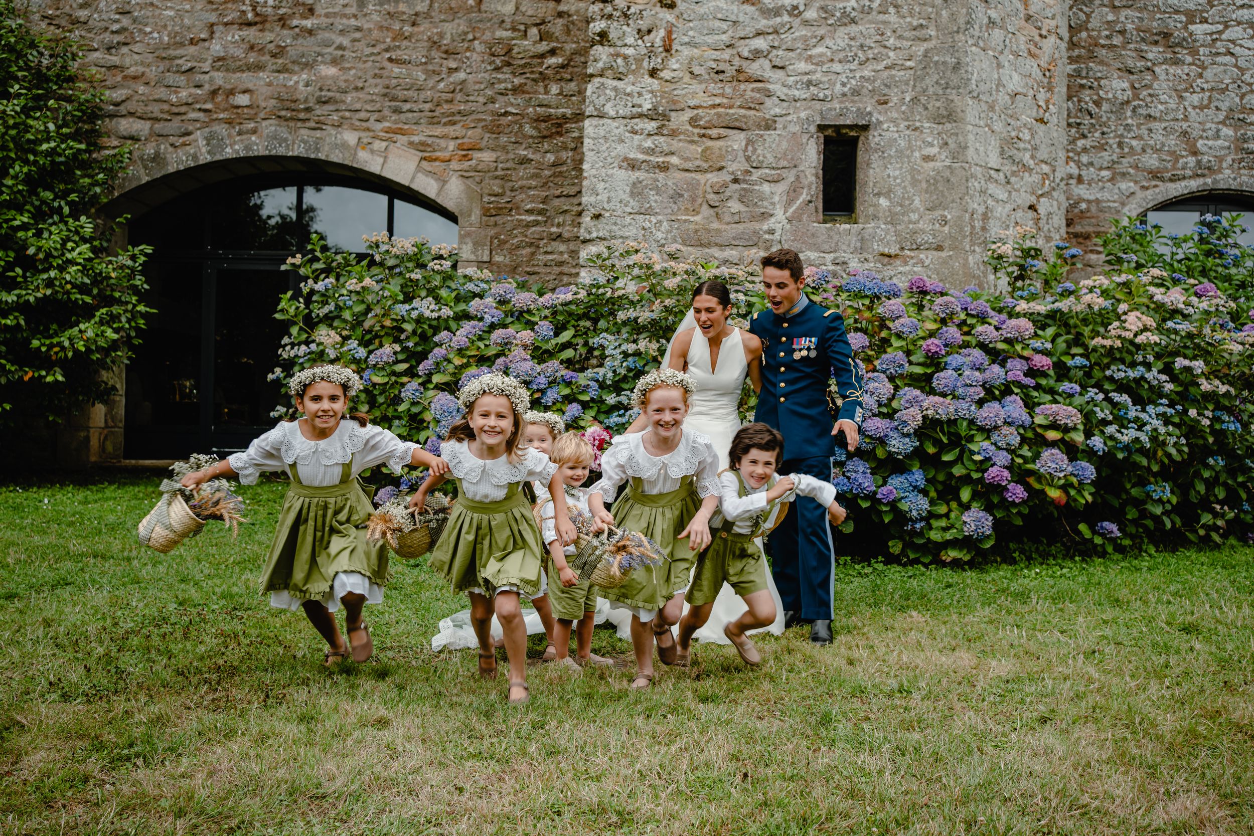 Wedding party poses playfully on grass in front of stone wall with purple hydrangeas.