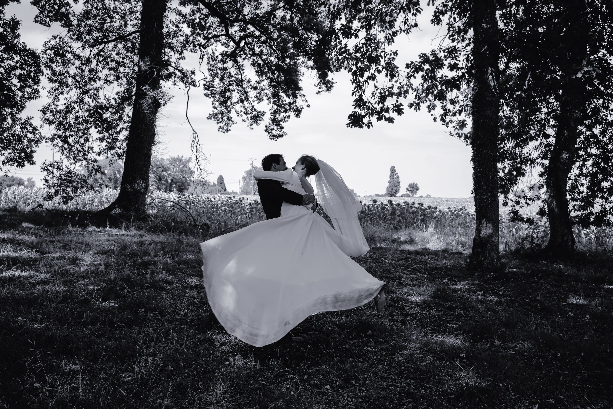 Artistic black and white photo of couple dancing on hillside under trees.