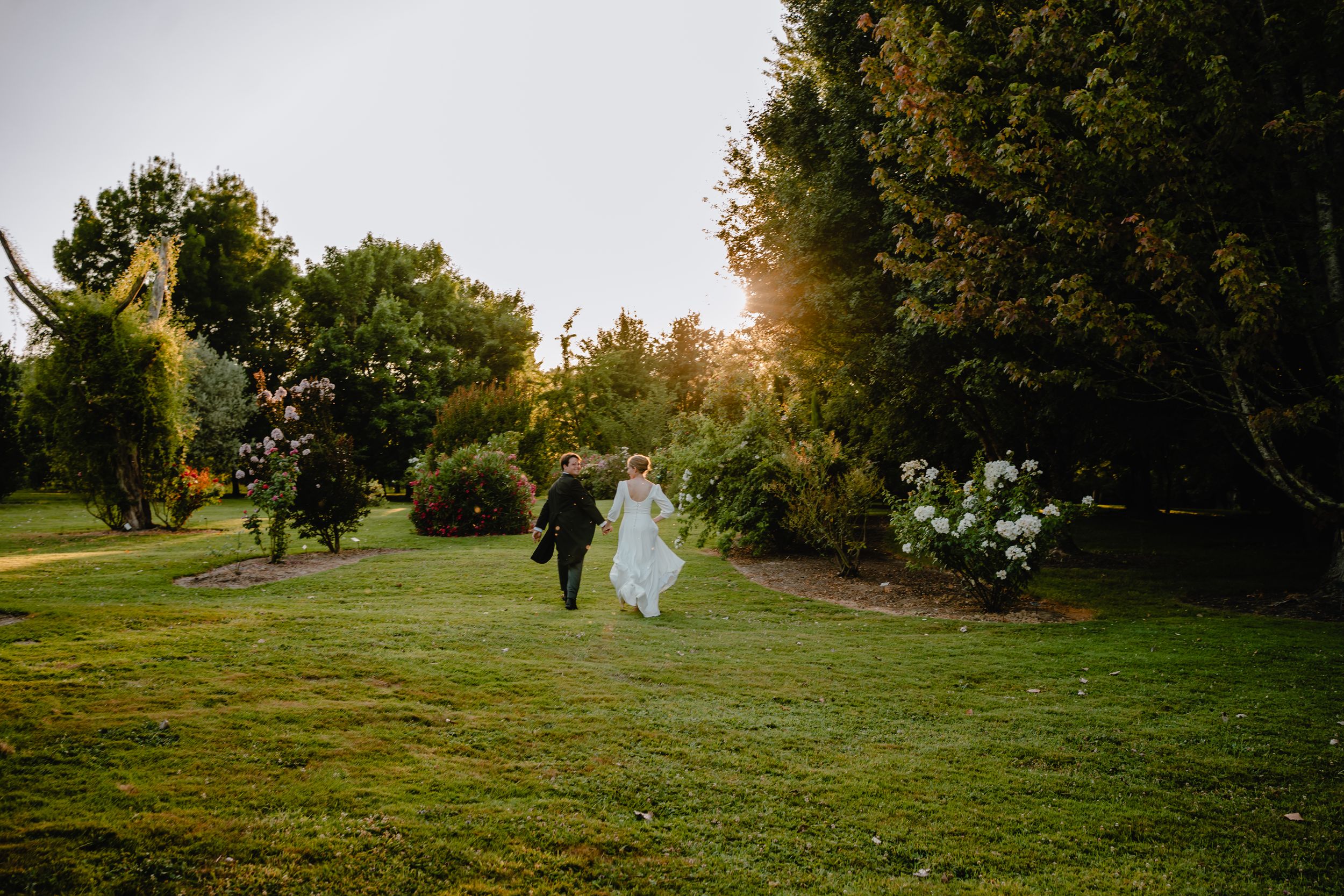 Wedding party walks across sunlit garden lawn at golden hour.