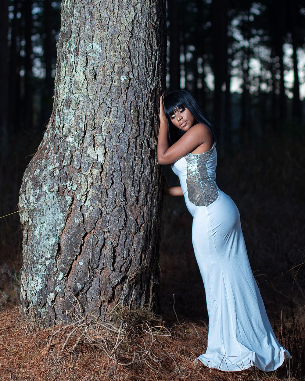 Artistic portrait in white flowing dress leaning against large tree trunk in forest setting.