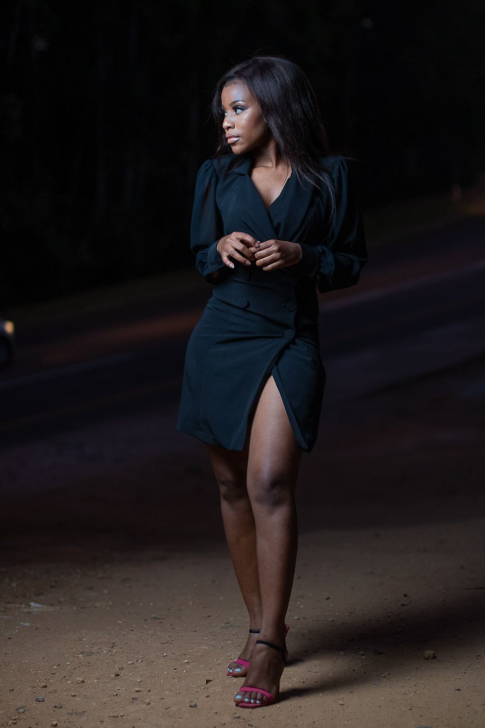 Woman in black dress with slit poses dramatically in evening lighting.