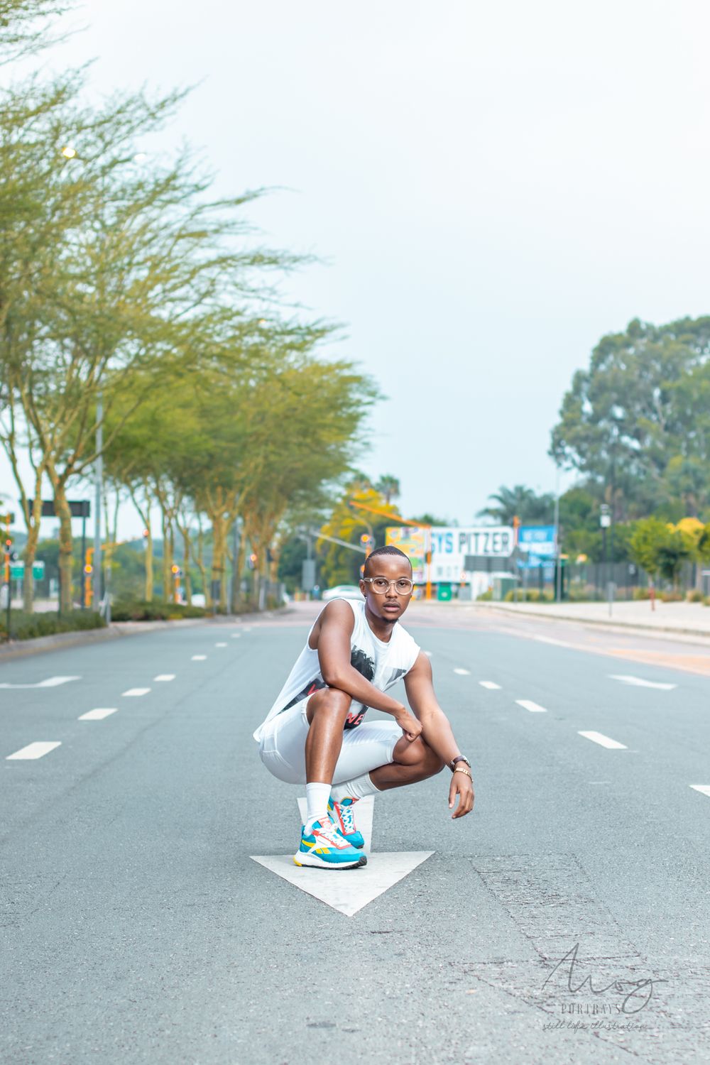 Urban fashion photo of person crouching on empty street wearing white outfit and blue sneakers.