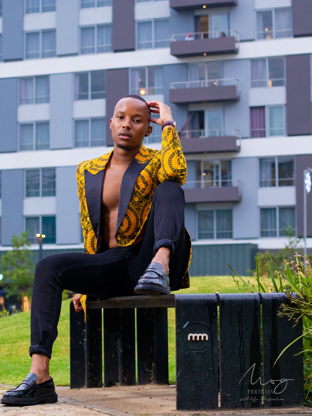 Model in black pants and gold patterned jacket poses on bench near modern apartment building.