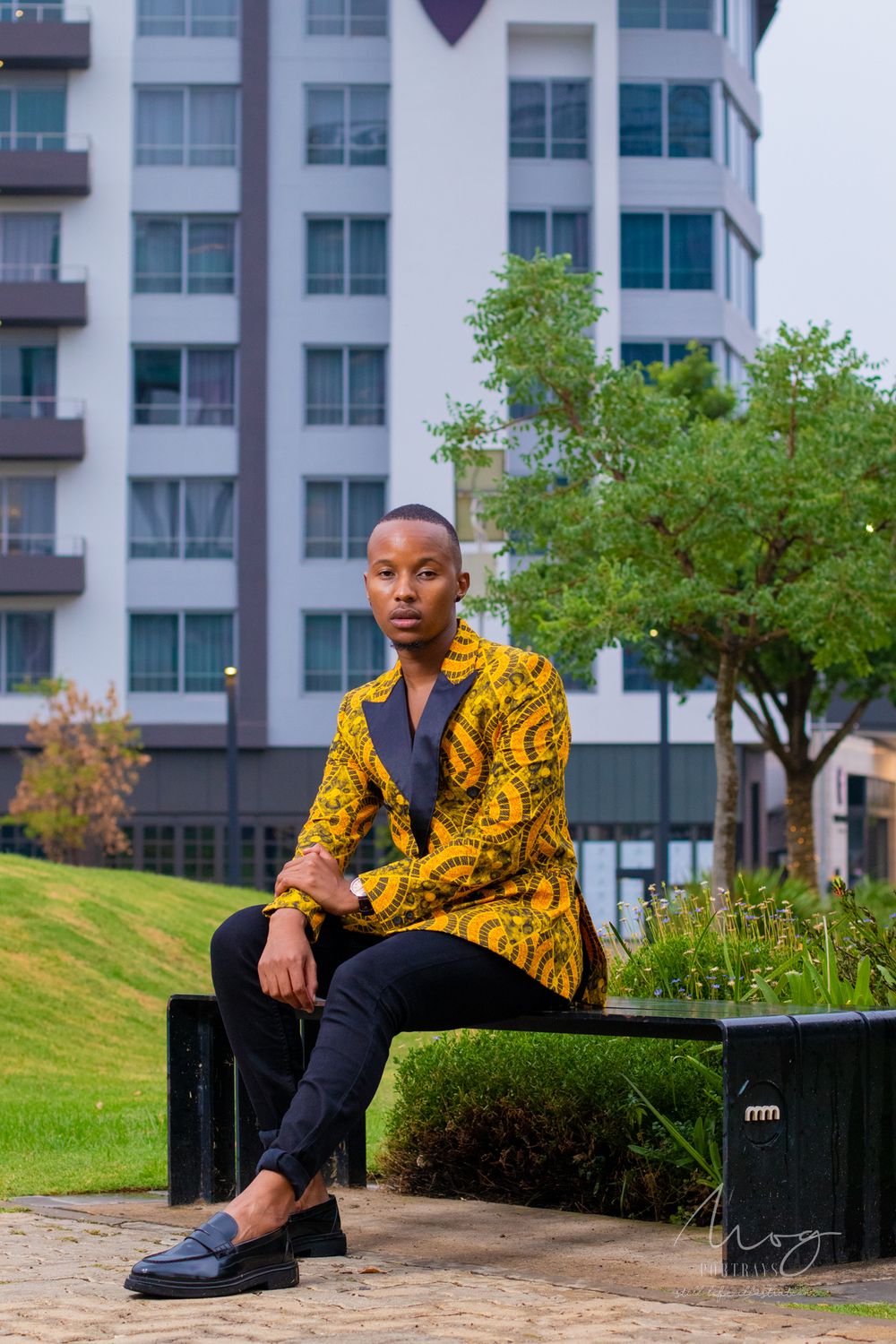 Model wearing yellow patterned blazer poses on bench near modern apartment building.