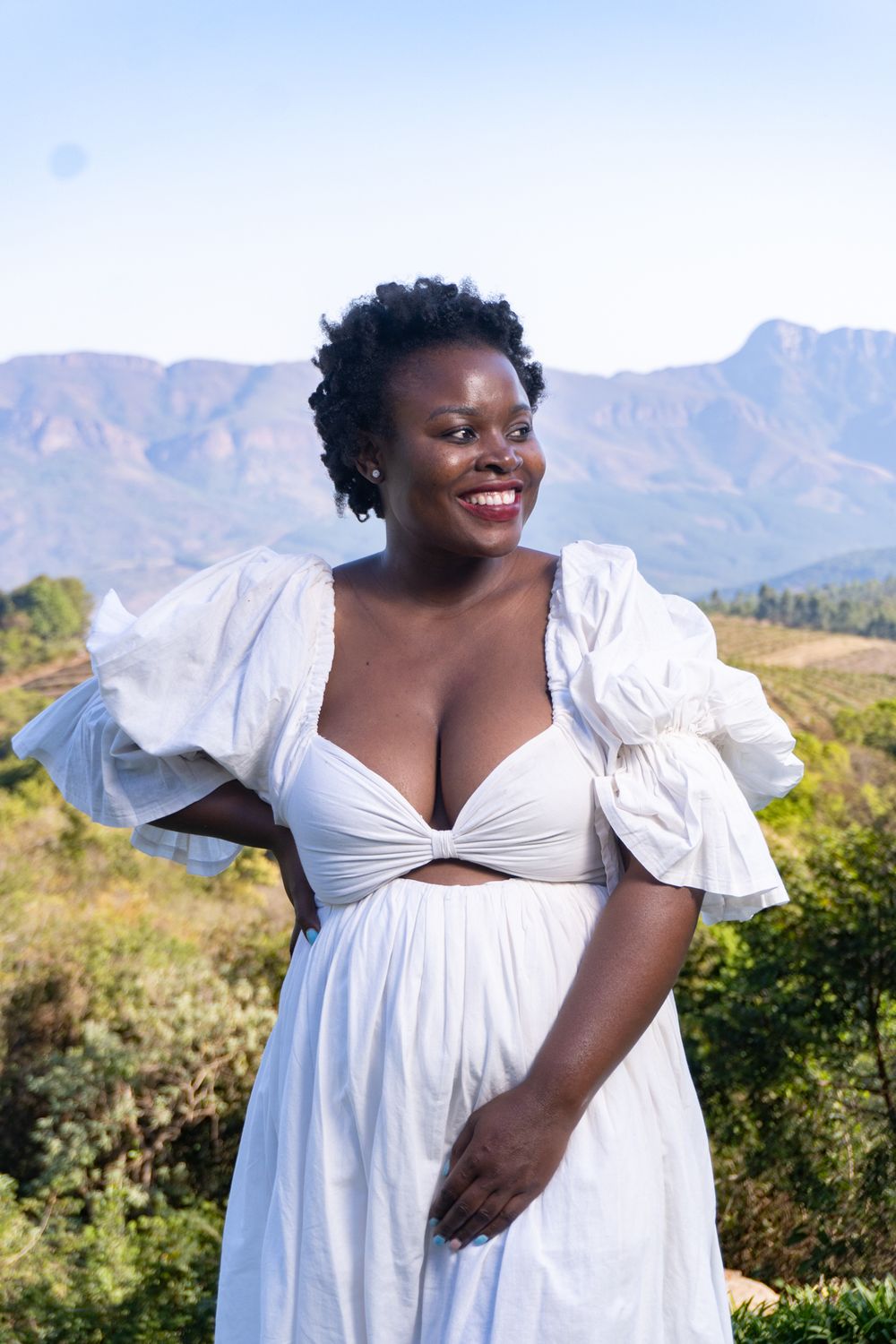 Person in elegant white dress poses against mountainous landscape backdrop.