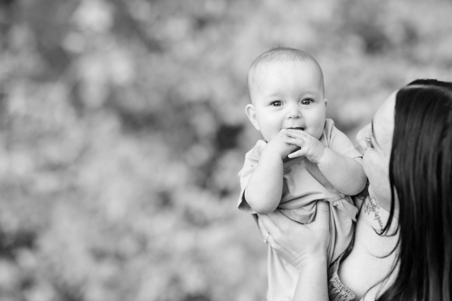 A black and white portrait of a baby being held against a blurred natural background.