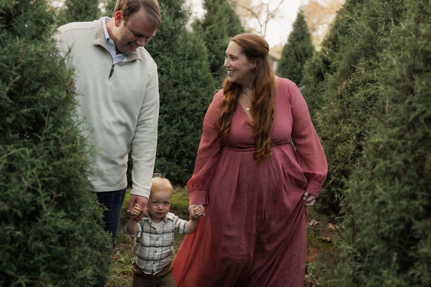 A family walks together between rows of evergreen Christmas trees at a tree farm while wearing winter attire.