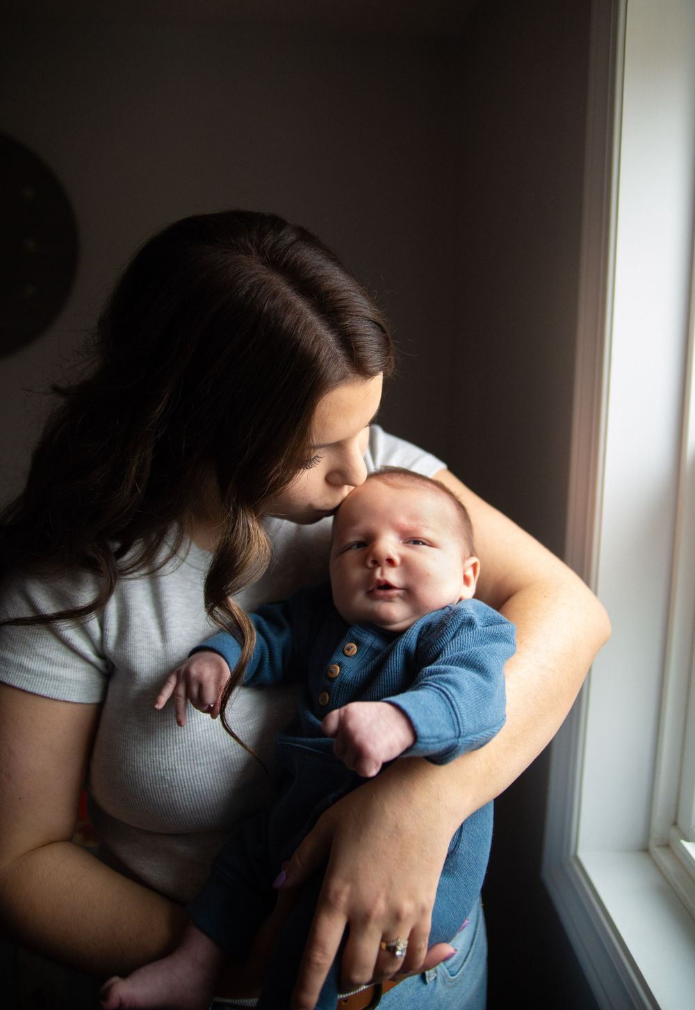 A tender moment by a window as a parent holds and kisses their baby wearing a blue outfit in natural light.