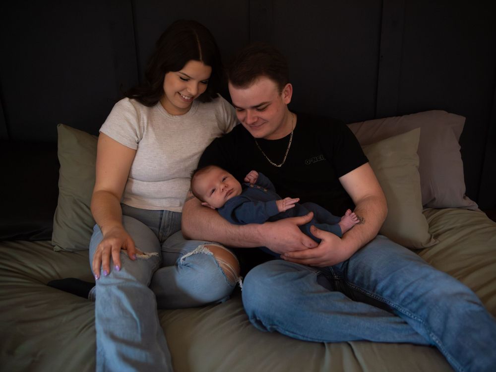 A couple sitting on a couch cuddles with their sleeping baby, creating an intimate family moment in dim lighting.
