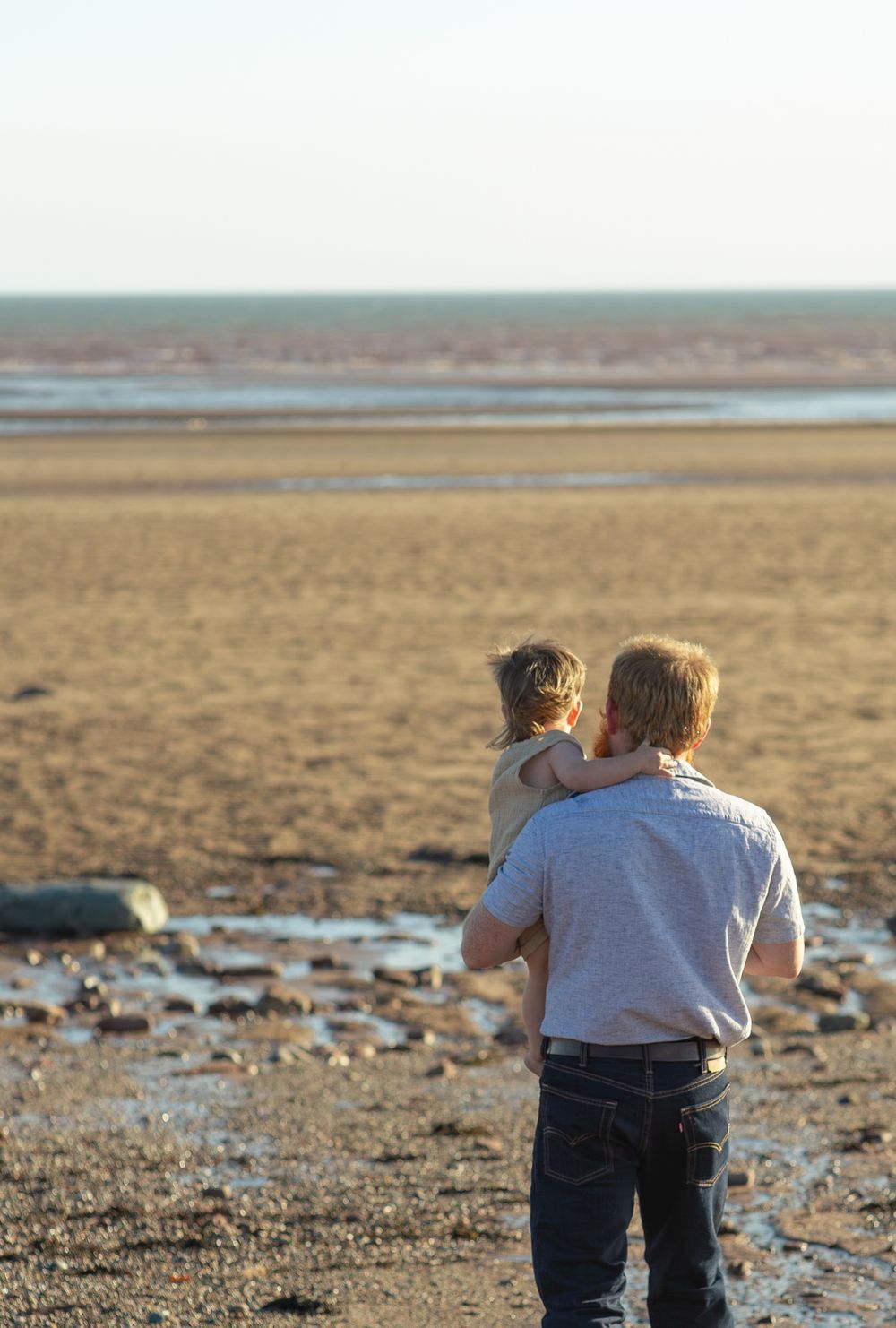 Two people crouch down to observe something along a rocky shoreline during sunset.