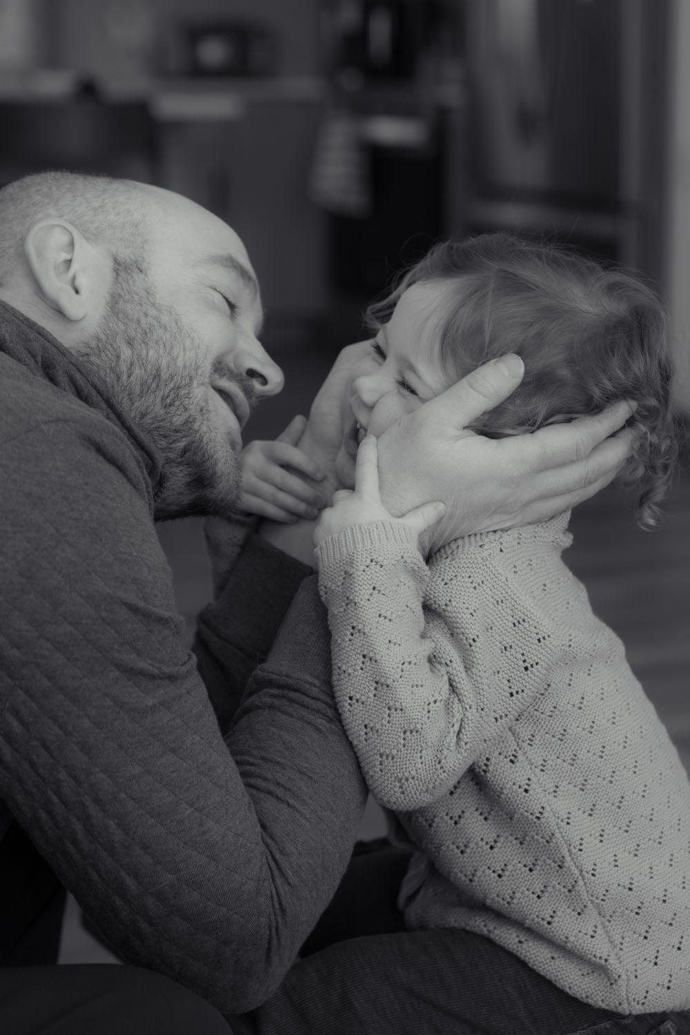 Black and white photo of a bearded father lovingly holding his toddler's face while they share a joyful moment.