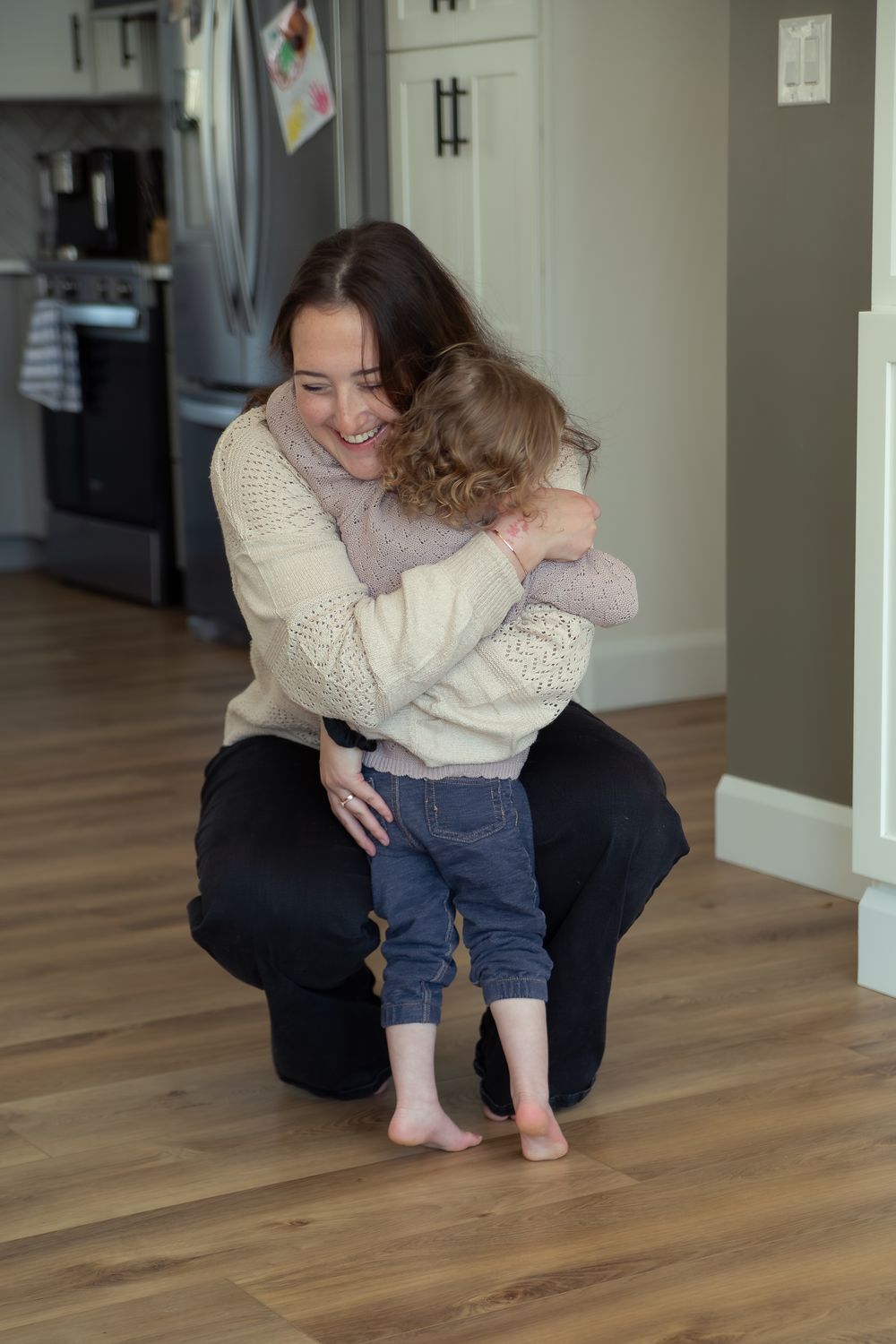 A smiling mother crouches to hug her toddler in a modern kitchen with hardwood floors.