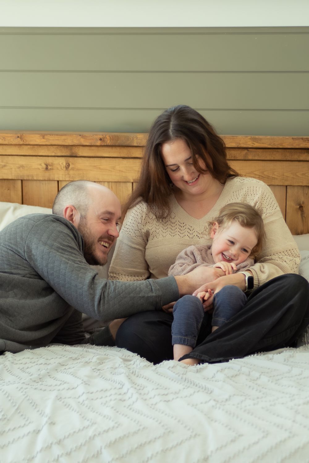 A happy family of three laughing together on a bed with a wooden headboard in a cozy bedroom setting.