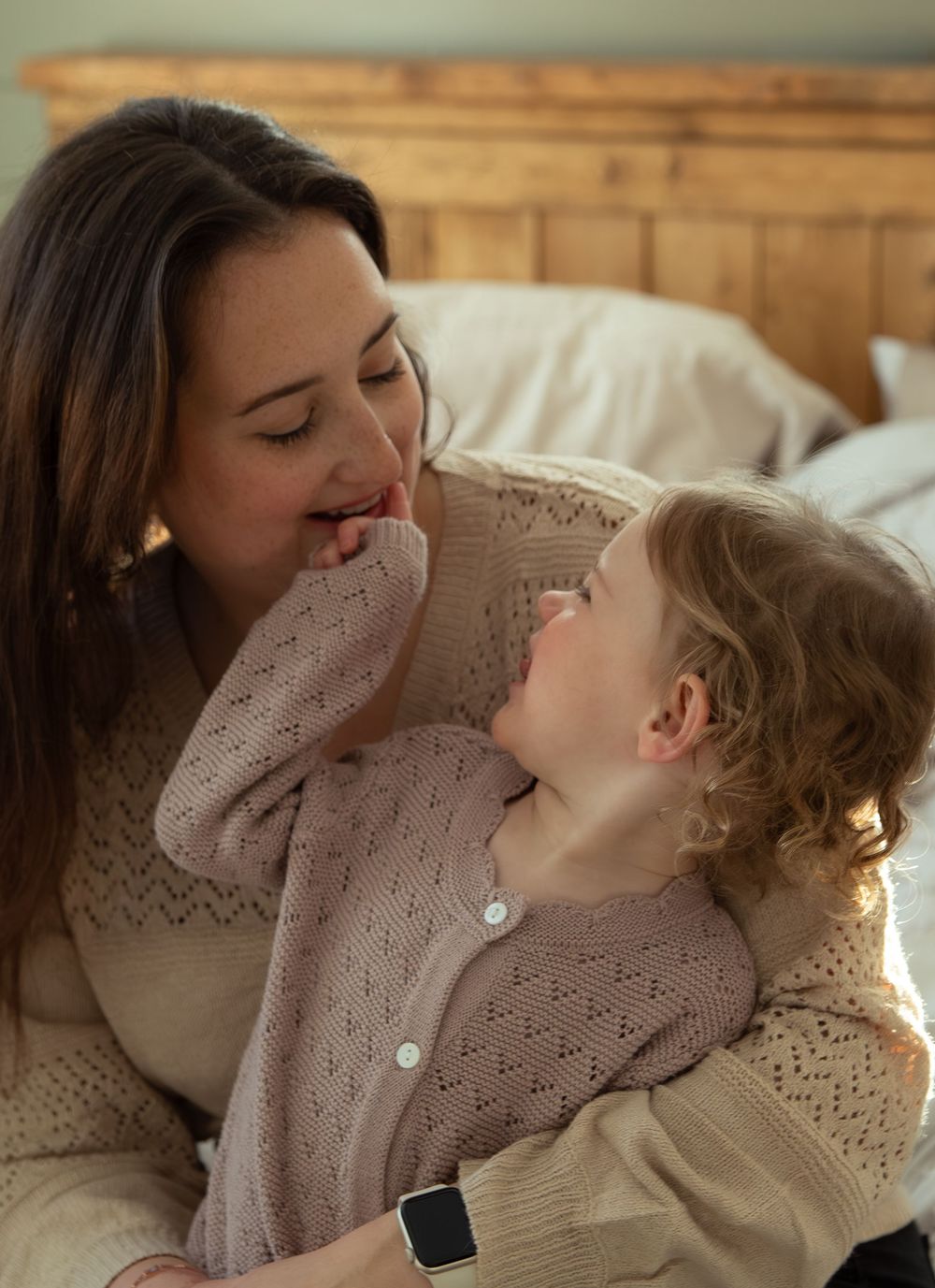A mother and toddler share a tender moment, both wearing cozy knit sweaters while sitting near a bed.