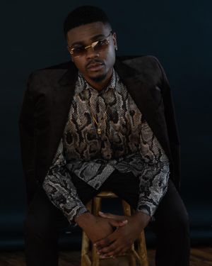 Portrait of person in snakeskin print shirt and black blazer seated on wooden chair.
