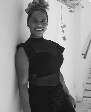 A black and white portrait photo showing a smiling figure in a stylish black dress with ruched details against a white wall.