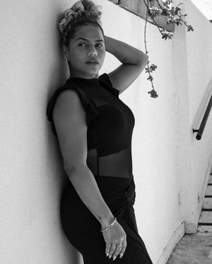 Black and white photo of someone in a sleeveless black outfit posing against a white wall with trailing vines.
