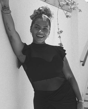 Black and white portrait of a beaming woman in dark clothing with upswept hair against a light wall.