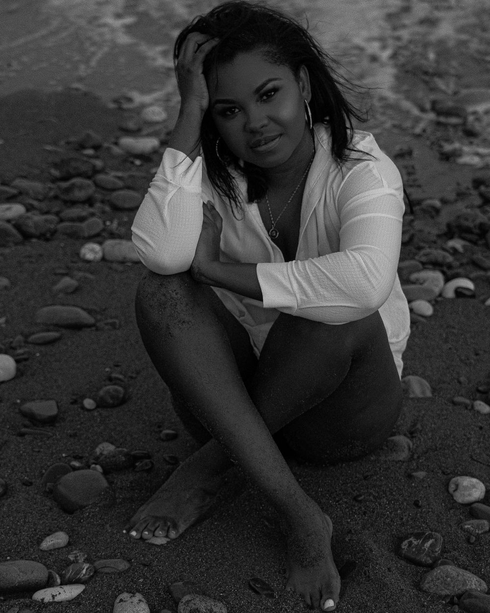Black and white photo of a woman sitting on a rocky beach, wearing a white shirt, posing gracefully.