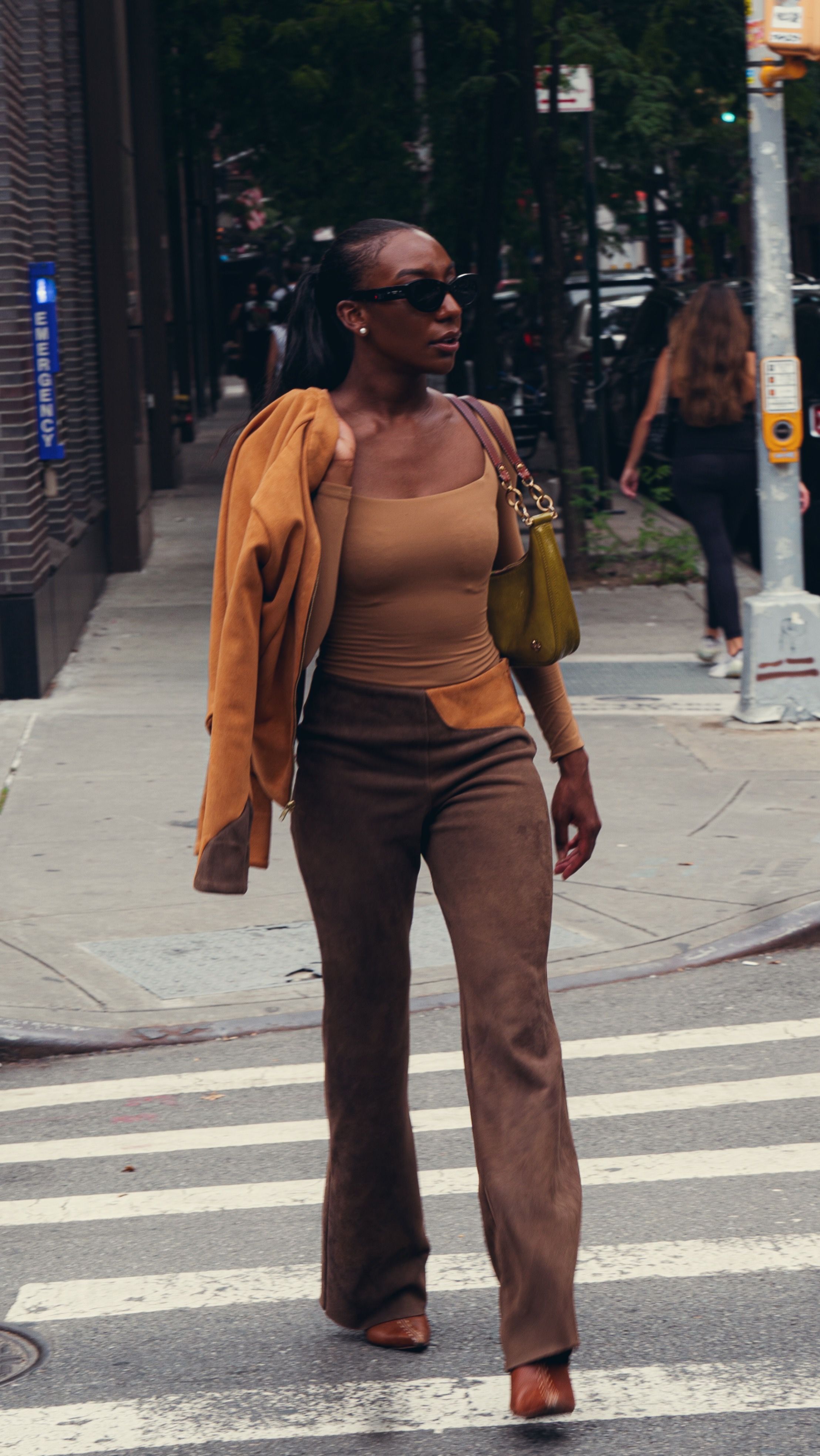 Stylish woman crossing street wearing tan tank top, brown pants, orange jacket and yellow handbag in urban setting.
