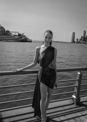 A stylish black dress silhouette stands on a pier overlooking a waterfront cityscape on a cloudy day.