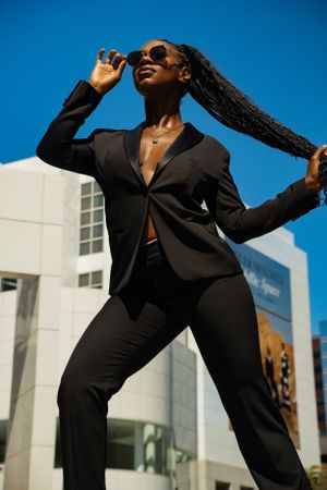 Stylish model in black blazer and pants poses confidently against modern building on sunny day.