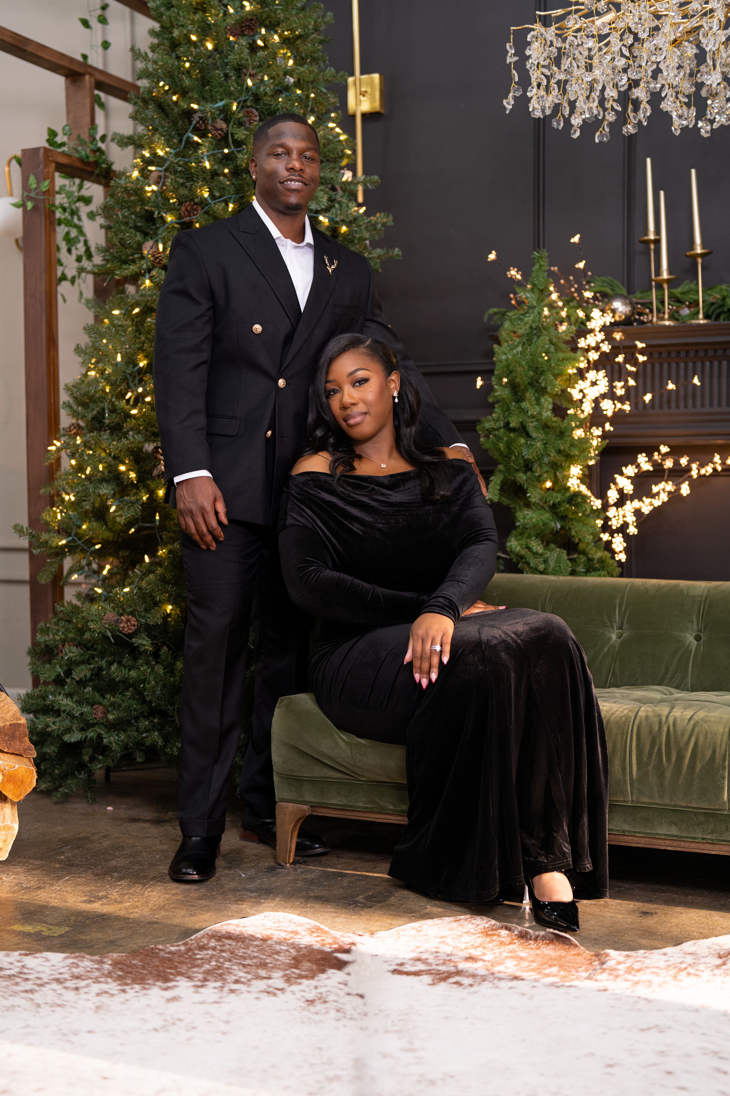 Elegant couple poses in formal black attire by a decorated Christmas tree and chandelier in an upscale holiday setting.