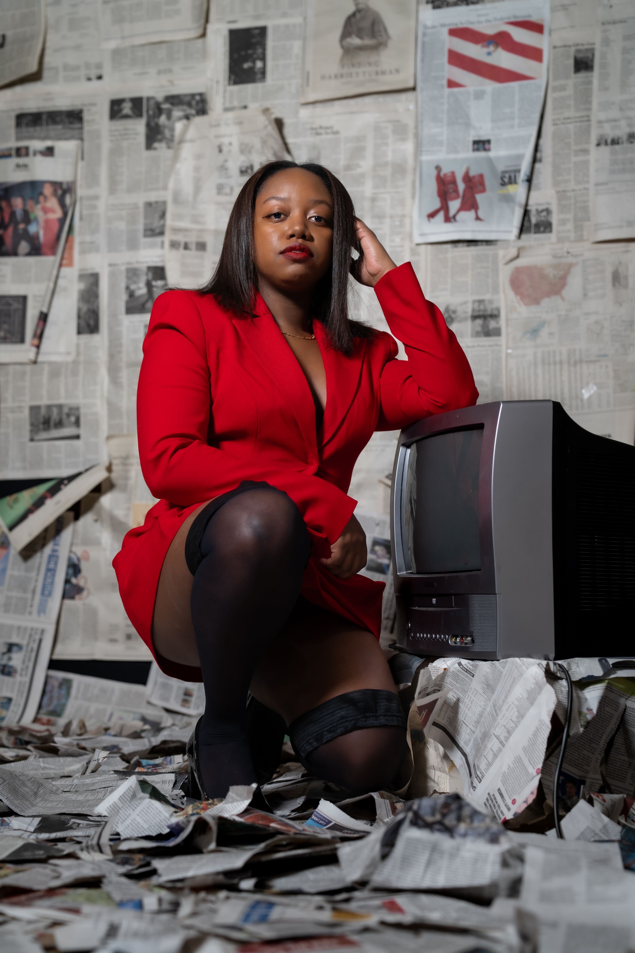 Woman in red blazer poses with vintage TV amid scattered newspapers and news-covered walls.