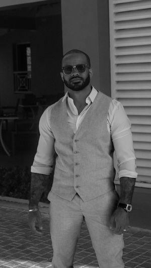 A well-dressed person in a light vest and rolled-up sleeves stands confidently while wearing sunglasses.