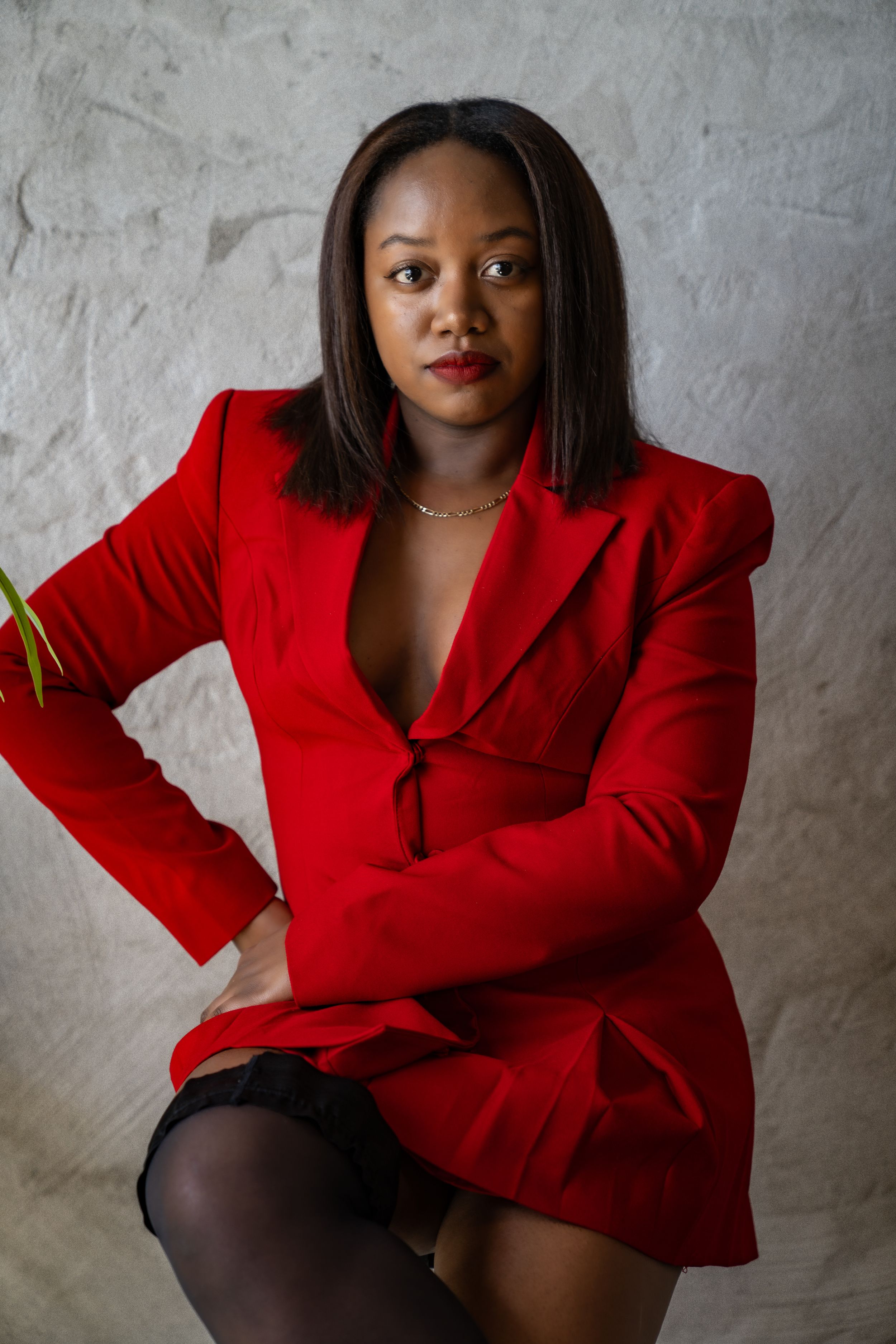 A confident woman in a red blazer sits against a gray wall, wearing black stockings and a gold necklace.